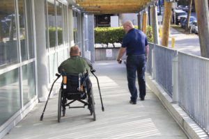 Two men move down a ramp side-by-side. One in a wheelchair, the other ambulatory.